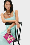 Woman holding the classic, high-end Fuchsia Champagne Problems Book Clutch, a fun and effortless statement piece, sitting stylishly on a black wooden chair.