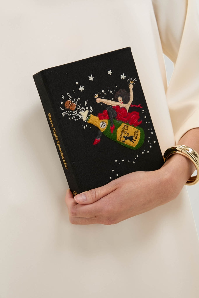 Woman holds the classic, high-end Black Starry Night Spectacular Book Clutch, featuring embroidered champagne and stars, epitomizing effortless, fun style.