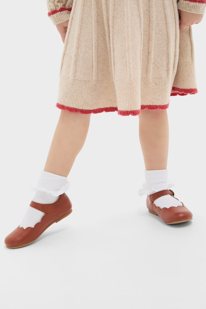 Child wearing the classic Sienna Skirt, showcasing its effortless pleats and fun red-trimmed scallop hem, paired with high-end brown Mary-Jane shoes and frilly socks.
