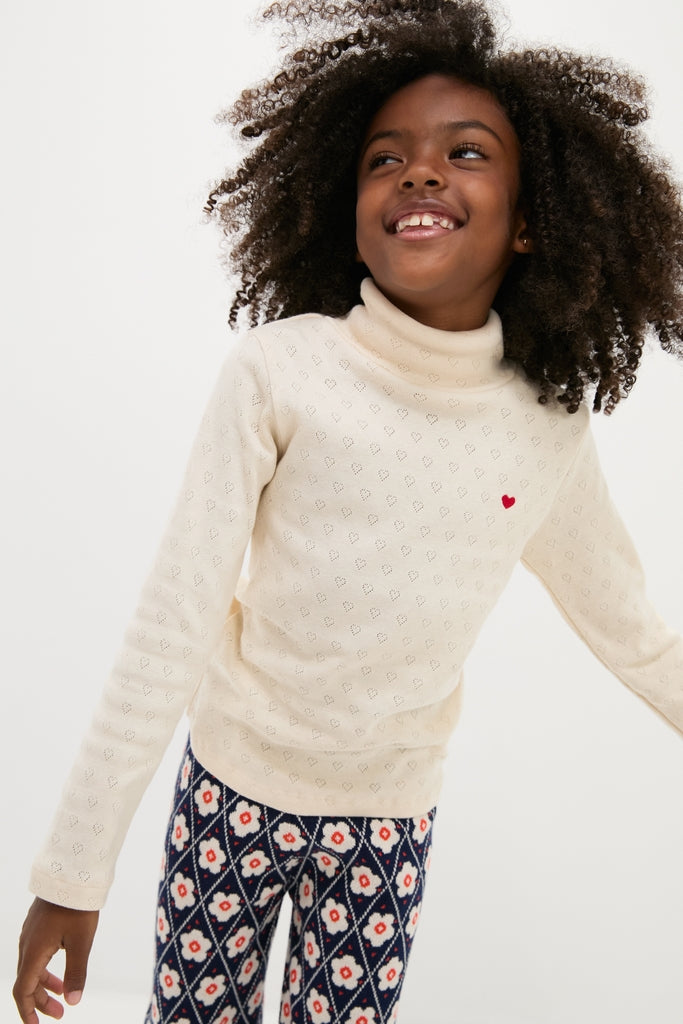 Child smiling in a classic Bebe Heart Turtleneck, effortlessly styled with navy leggings, showcasing fun heart embroidery and high-end organic cotton comfort.
