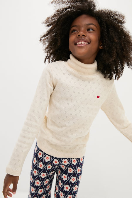 Child smiling in a classic Bebe Heart Turtleneck, effortlessly styled with navy leggings, showcasing fun heart embroidery and high-end organic cotton comfort.