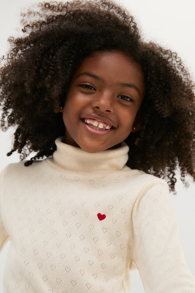 Young girl in a classic Bebe Heart Turtleneck, featuring an effortless, fun heart pattern and embroidery, embodying high-end style with a snug, comfortable fit.