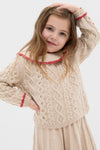 Young girl wearing the Sienna Sweater, a classic and effortless cream cable-knit with ruffled collar and pink trim, posing playfully in a fun, high-end style.