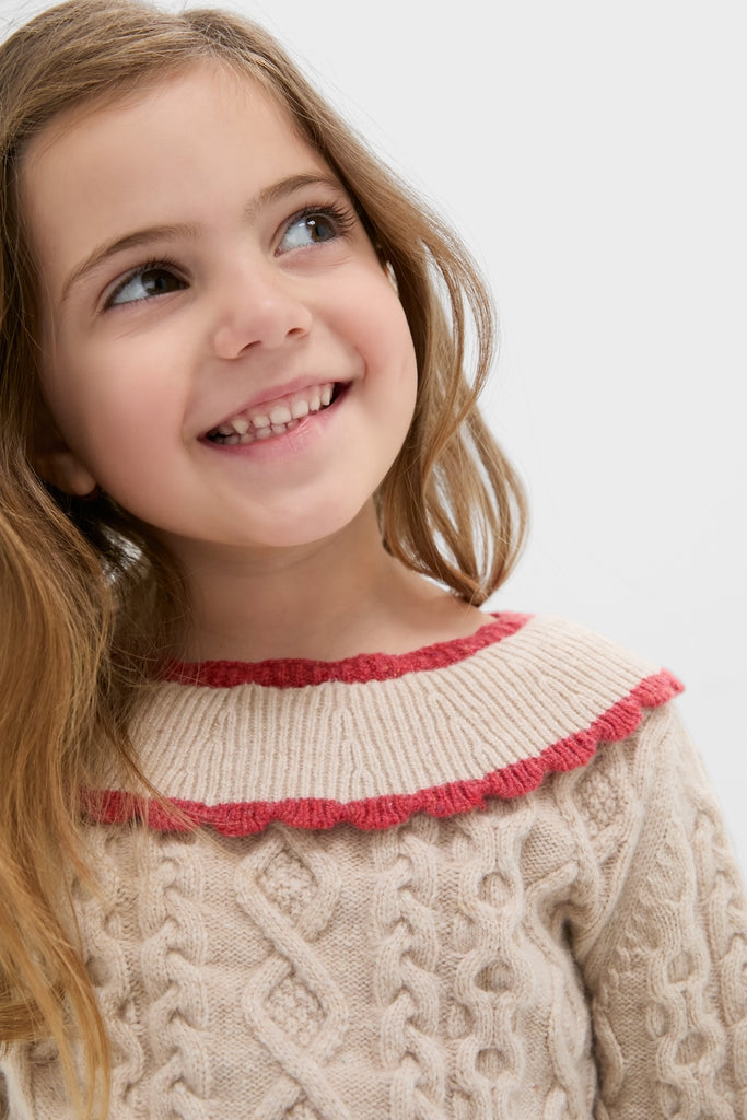 Young girl in a cream Sienna Sweater with ruffled neckline, showcasing classic cable-knit design and effortless style, perfect for fun, high-end cold-weather fashion.