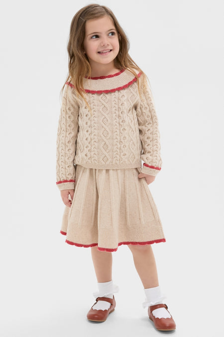 Young girl in Sienna Sweater: classic cable-knit with red-trimmed ruffles, pleated skirt, embodying effortless, high-end style for fun, chic cold-weather fashion.