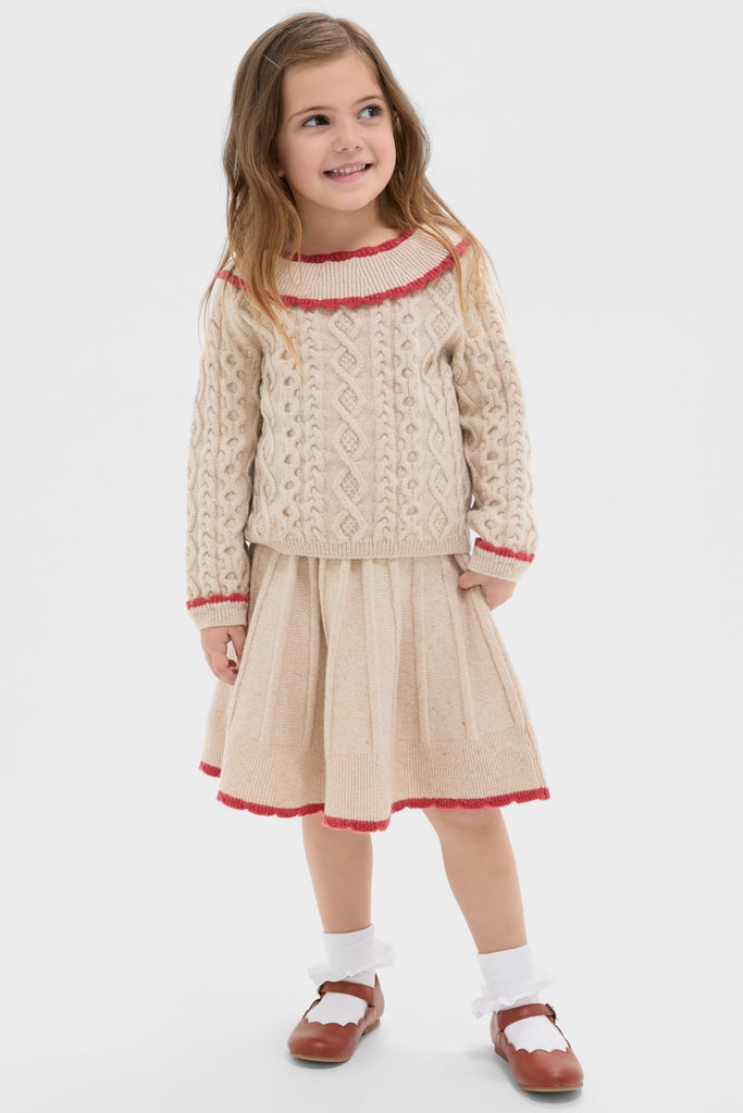 Young girl in Sienna Sweater: classic cable-knit with red-trimmed ruffles, pleated skirt, embodying effortless, high-end style for fun, chic cold-weather fashion.