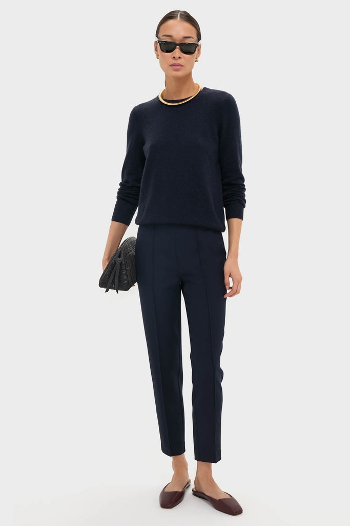 Woman in classic Navy Audrey Pants, styled effortlessly with a dark sweater and burgundy flats, accessorized with gold jewelry and a quilted clutch for a high-end look.