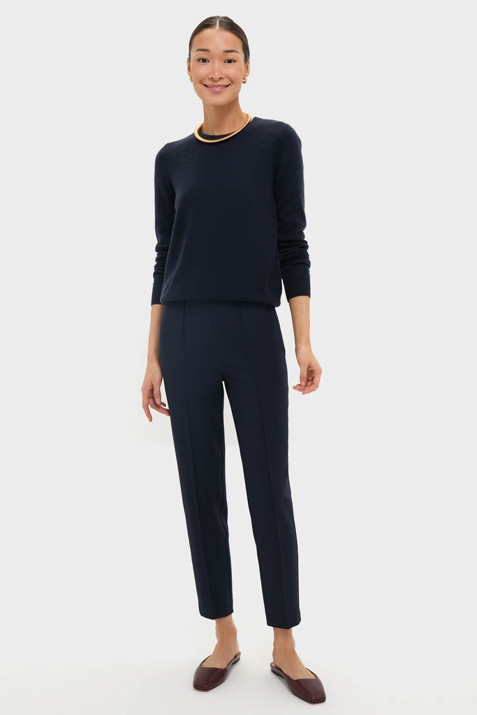 Woman in Navy Audrey Pants, classic and effortless, styled with a navy sweater and burgundy mules. Features pinched seams and zipper closure for a high-end, fun look.