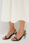 Model wearing Maplewood Aurora Sling Mid Heels, black strappy sandals with buckle details, paired with a white skirt. Classic, effortless, and high-end style.