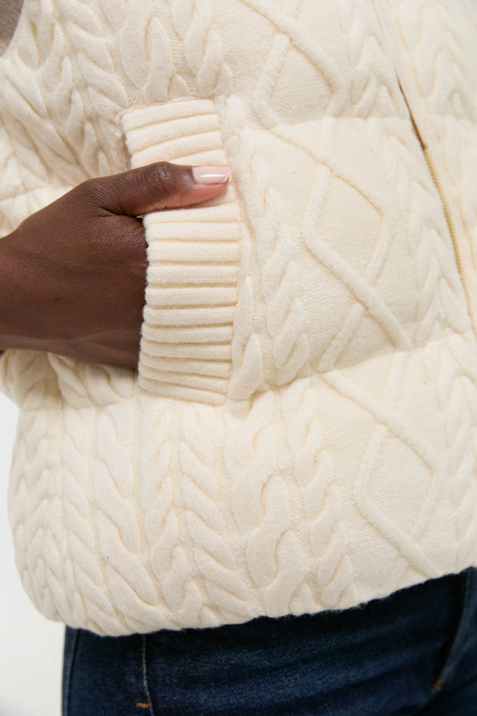 Close-up of a hand in the Egret Irina Cable Knit Gilet's pocket, showcasing classic cable-knit pattern with rib-knit trim, exuding effortless high-end style.