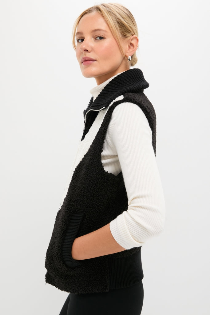 Young woman models the Black Caspian Sherpa Gilet, a classic, high-end, zip-up vest with side pockets, over a ribbed white top, exuding an effortless, fun style.