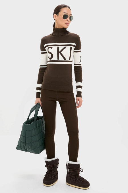 Model wearing Chocolate Schild Sweater with SKI design, styled with leggings and fur-trimmed boots, holding a quilted tote. An effortless, high-end look for winter fun.