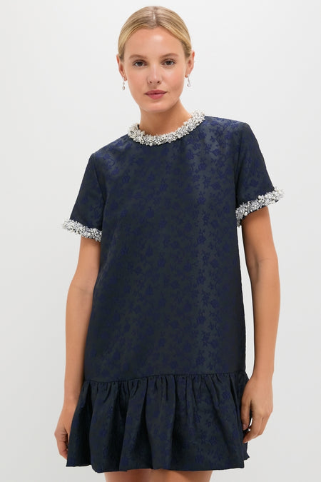 Model wearing the Midnight Alison Short Sleeve Mini Dress, a classic, high-end navy-blue piece with effortless floral patterns, faux-pearl details, and a fun, ruffled hem.