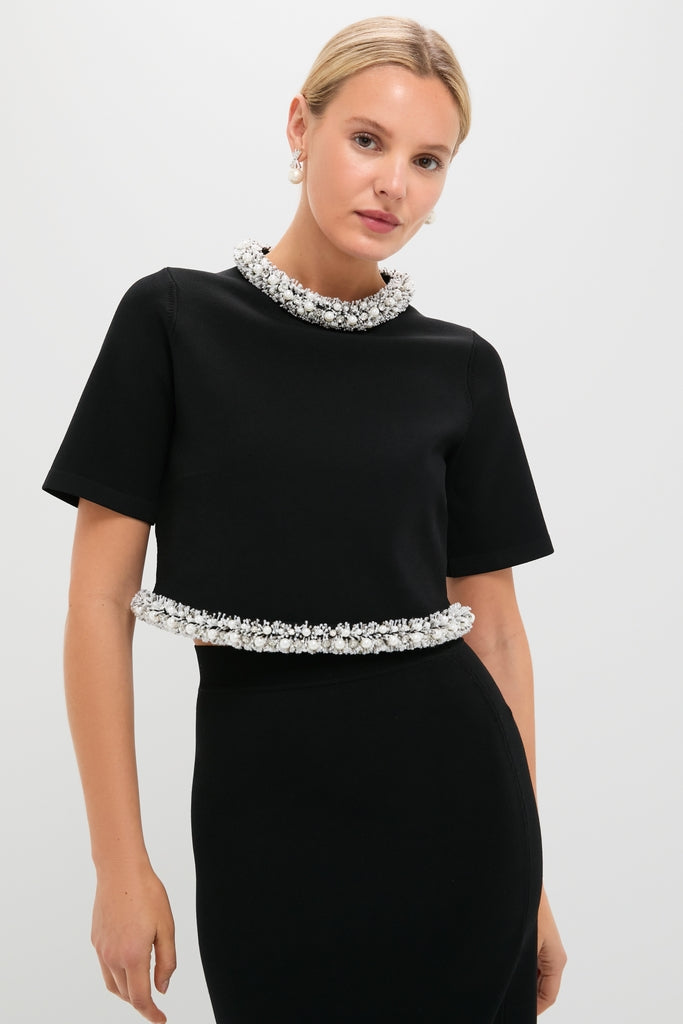 Woman models Black Ozelle Short Sleeve Crewneck Top, featuring a classic, effortless design with fun white bead embellishments at the neckline and hem, paired with a black skirt.