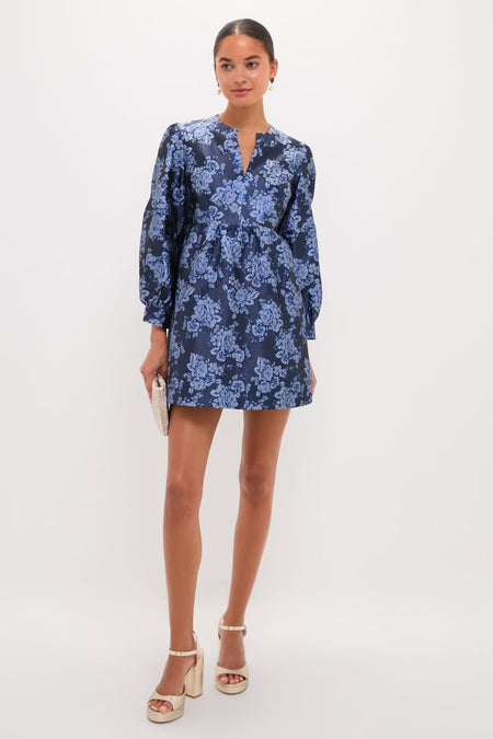 Young woman models the Blue Jacquard Cressida Mini Dress with a classic floral pattern, effortless V-notch neckline, and fun, high-end style, holding a metallic clutch.