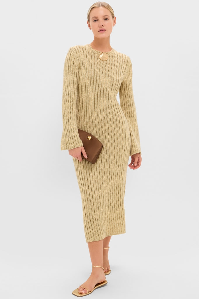 Woman models the Gold Knit Amanyara Maxi Dress, a classic rib-knit in warm beige, with long sleeves, round neckline, and mid-calf length, paired effortlessly with a brown clutch and gold sandals.