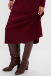 Person wearing the Oxblood Knit Conrad Midi Swing Dress, a classic ribbed design, paired with knee-high, brown leather heeled boots, embodying an effortless and high-end style.