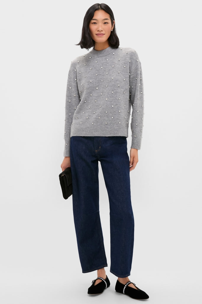 Woman in Charcoal Diamonte Aura Sweater with pearl embellishments, dark indigo jeans, and pearl-studded flats, holding a black clutch. Effortlessly classic and high-end style.