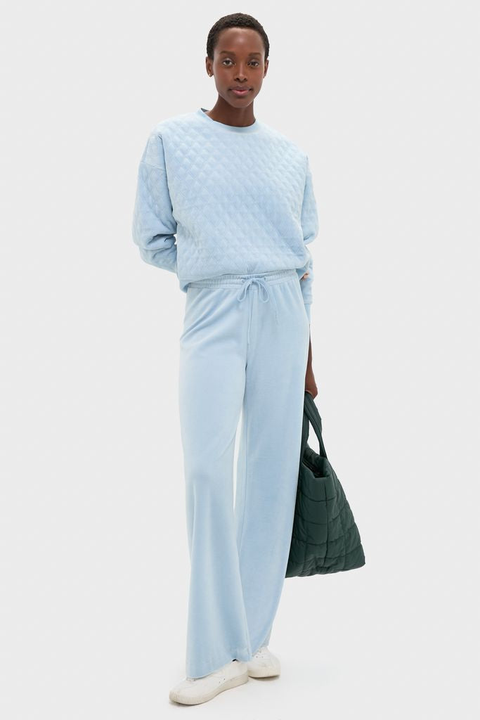 Woman models Blue Fog Velour Jane Pant, showcasing classic wide-leg, high-rise style with effortless fit, paired with a quilted tote, embodying high-end, fun loungewear.