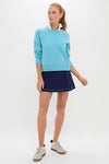 Young woman wearing the classic Bright Blue Washable Cashmere Malcolm Crewneck with raglan sleeves, layered over a white shirt, paired with a navy scalloped skort.