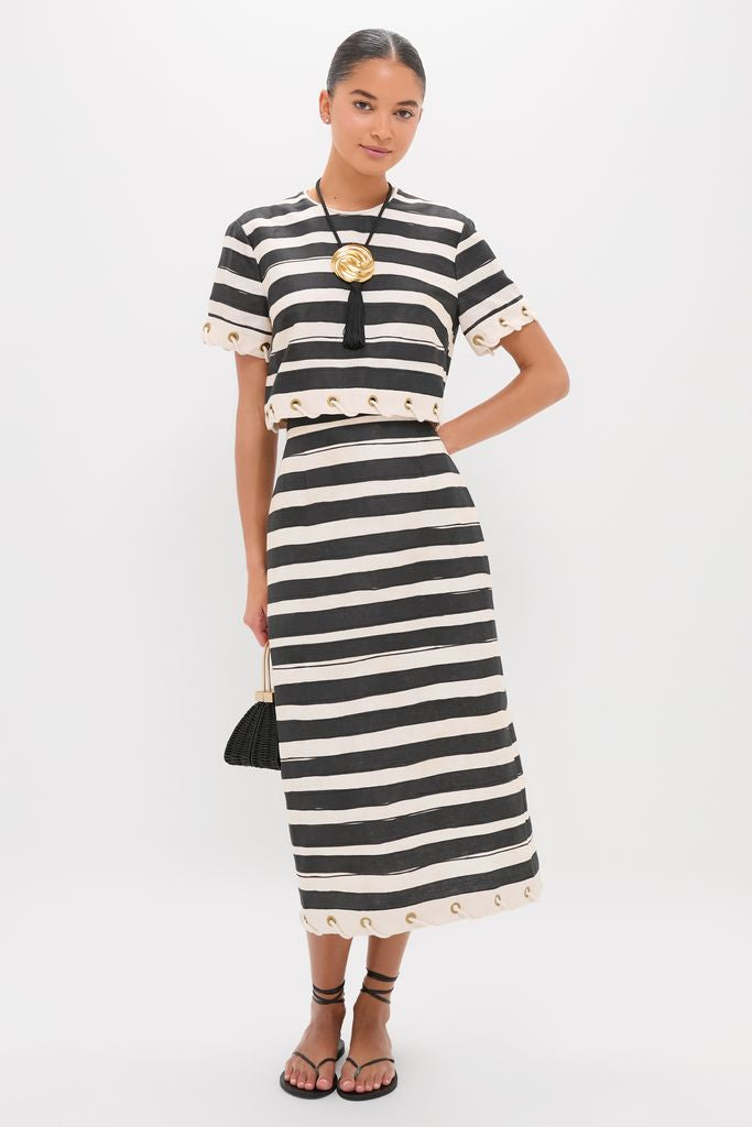 Woman models the Black Stripe Story Top, a classic, effortless knit with gold-tone grommets, paired with a matching skirt, statement necklace, and woven handbag for a high-end, fun summer look.