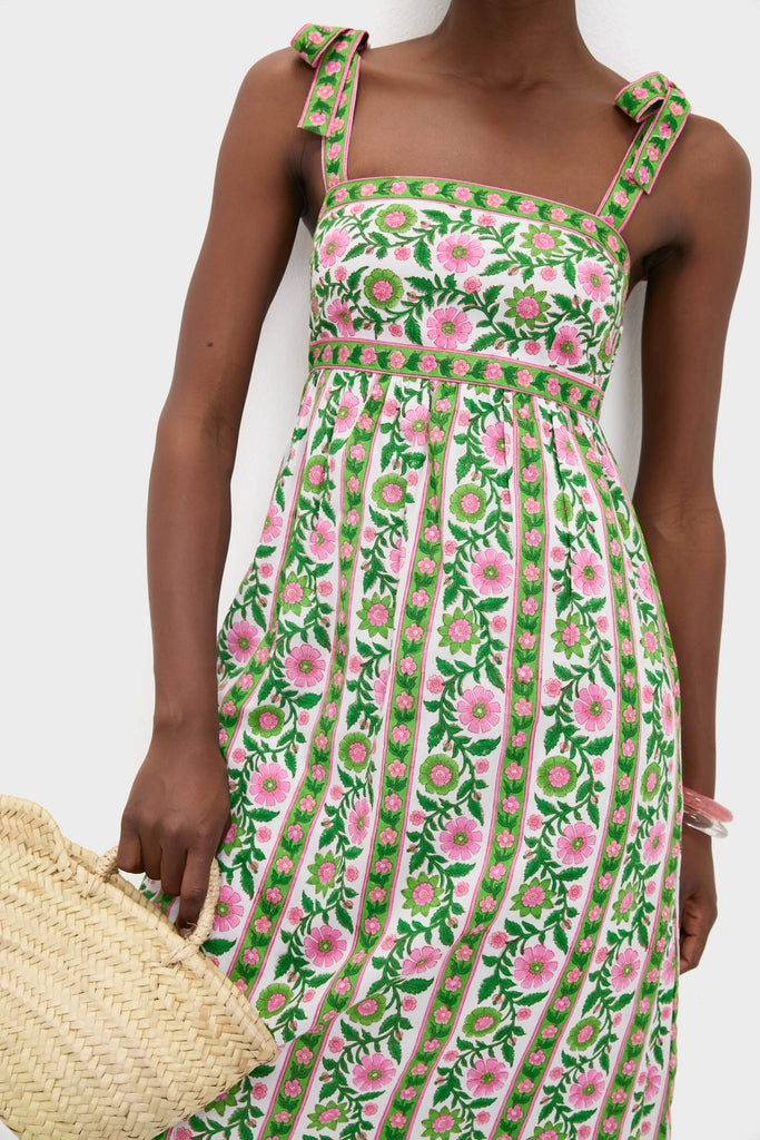 Model wearing the classic, high-end Emerald Border Mykonos Dress with pink floral bands, slim bow-tied straps, and a gathered skirt, paired with a straw tote and clear bangle.