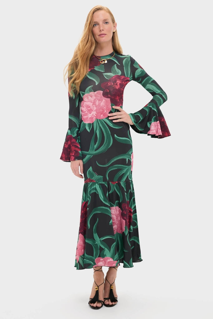 Model wearing the classic Peony Alba Dress, an effortless black maxi with vibrant floral patterns, flared sleeves, and a flounced hem, paired with high-end black heels and a gold necklace.