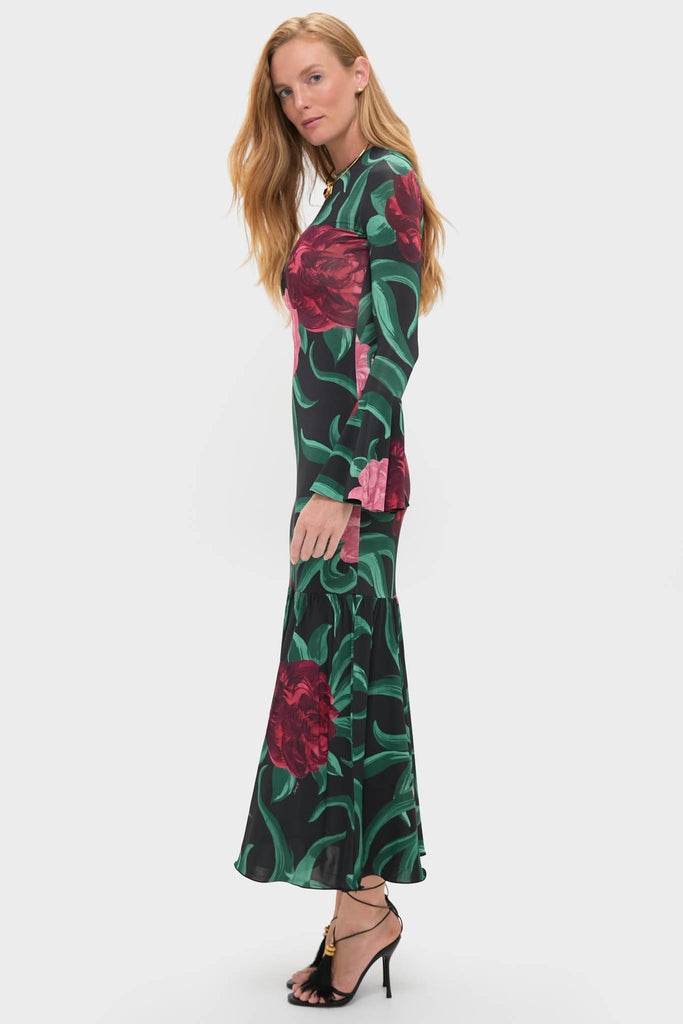 Model in profile wearing the Peony Alba Dress, a high-end, classic black gown with vibrant floral print, long sleeves, and a mermaid hem, styled with strappy sandals.