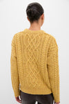 Woman in mustard Sunflower Madelyn Sweater with intricate cable knit, showcasing classic, effortless style with high-end appeal. Dark hair in bun, gold hoop earrings visible.