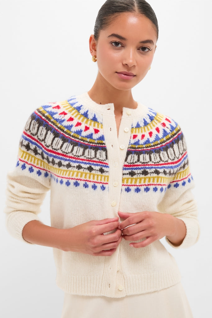 White Amina Fairisle Sweater modeled in a studio, showcasing its classic, multicolored yoke. The young woman fastens buttons, embodying an effortless, fun, high-end style.