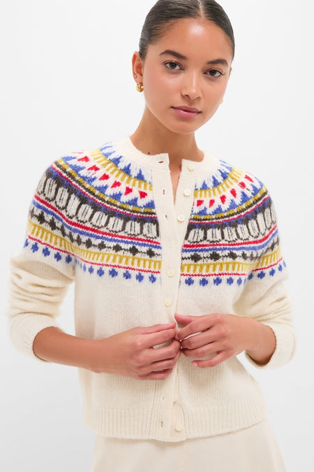 White Amina Fairisle Sweater modeled in a studio, showcasing its classic, multicolored yoke. The young woman fastens buttons, embodying an effortless, fun, high-end style.
