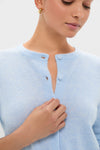 Woman wearing an Ice Blue Cashmere Riley Cardigan, showcasing its classic and effortless style. The cardigan is soft-knit with top buttons undone, complemented by small gold hoop earrings.