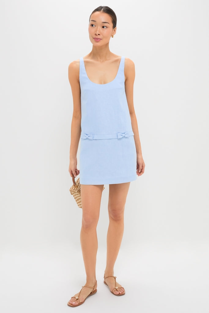 Young woman in the Blue Harper Mini Dress, featuring a scoop neck and decorative waist bows, paired with tan sandals and a woven straw handbag for an effortless, high-end look.