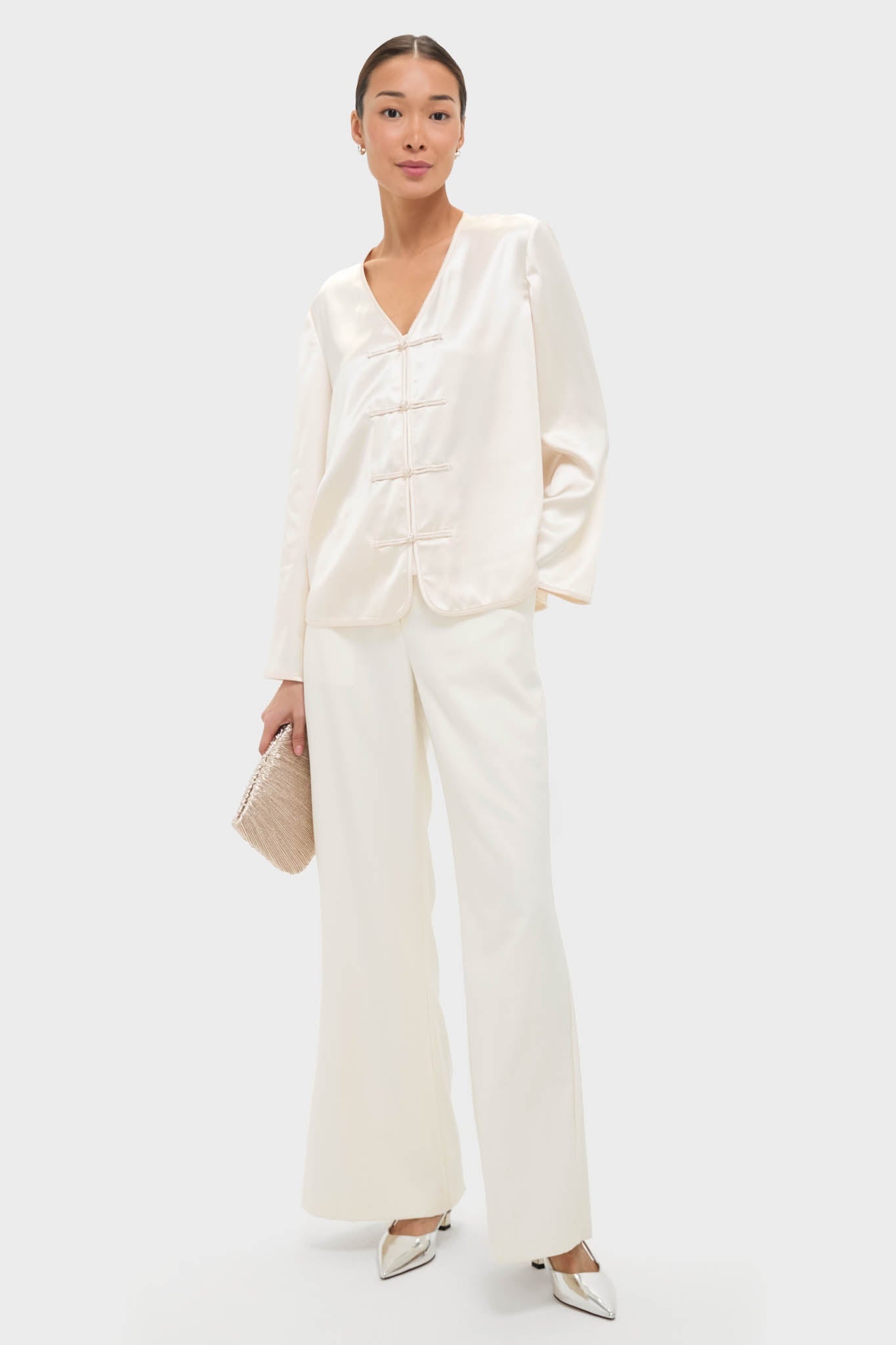 Cream Satin Tatiana Blouse with classic V-neck and chic frog closures styled effortlessly with matching trousers, metallic shoes, and a woven clutch for a high-end, fun look.