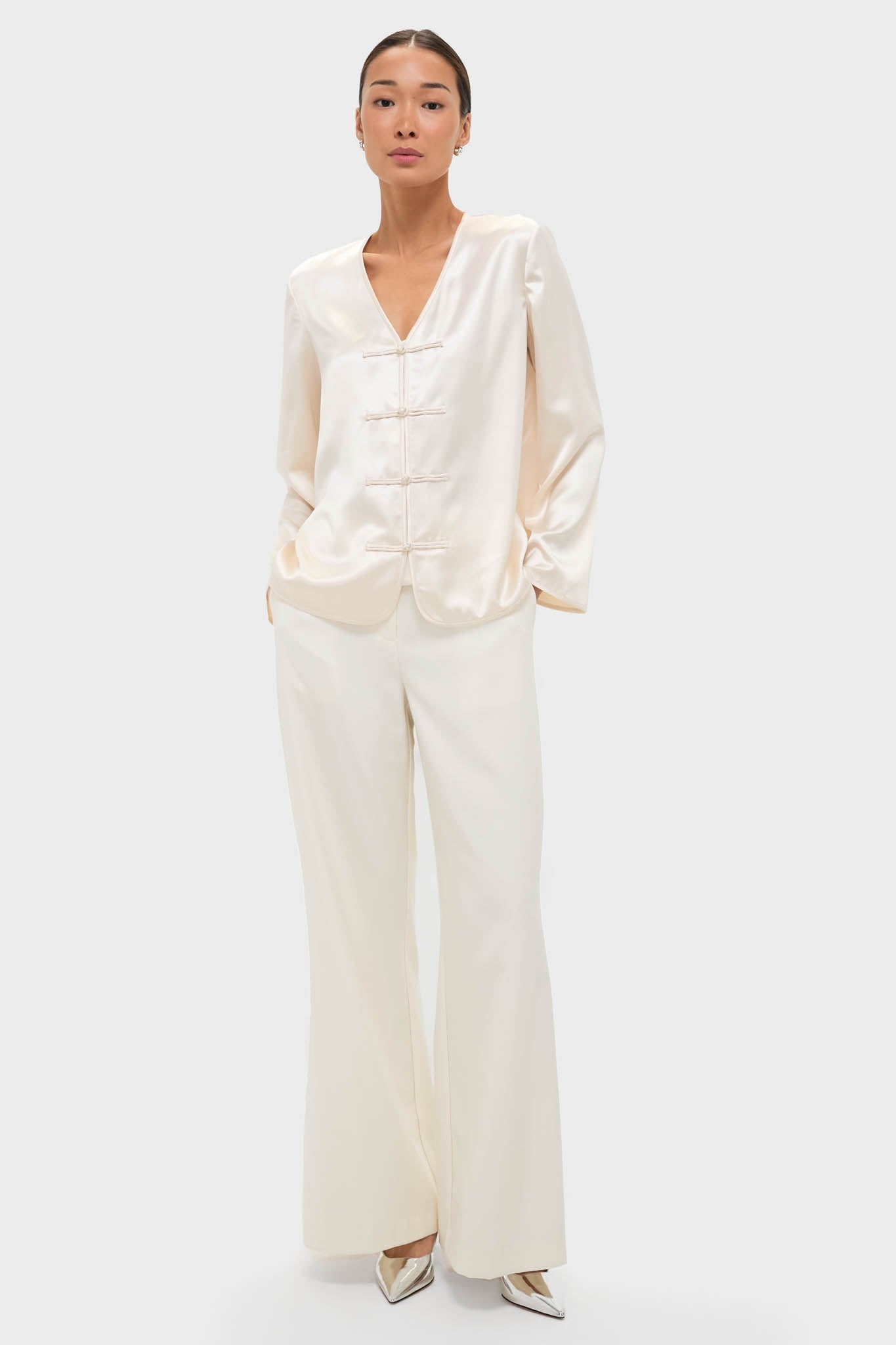 Model wearing the classic Cream Satin Tatiana Blouse with frog closures and a relaxed fit, paired with high-waisted trousers and metallic heels for an effortless, high-end look.