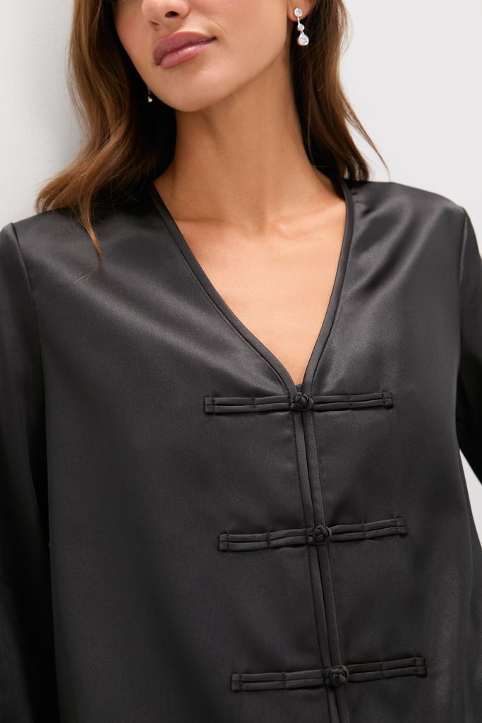 Woman in Black Satin Tatiana Blouse with classic V-neck and frog closures, showcasing effortless style. Pair with accessories for a fun, high-end look.