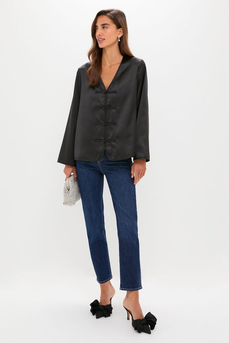 Woman models the Black Satin Tatiana Blouse with V-neck and frog closures, paired with jeans, clutch, and heels for an effortless, high-end, day-to-night chic look.