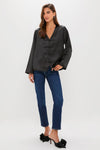 Woman wearing the classic Black Satin Tatiana Blouse with V-neck and frog closures, paired with effortless mid-blue jeans and fun black mule pumps for a high-end look.