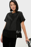 Woman wearing Black Dupioni Chadwick Tassel Top with tassel hem, classic round neck, and relaxed fit, leaning on chair, exuding an effortless, high-end style.