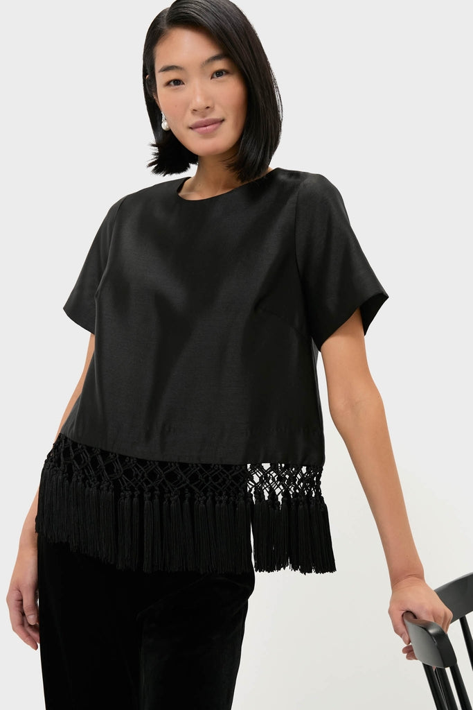 Woman wearing Black Dupioni Chadwick Tassel Top with tassel hem, classic round neck, and relaxed fit, leaning on chair, exuding an effortless, high-end style.