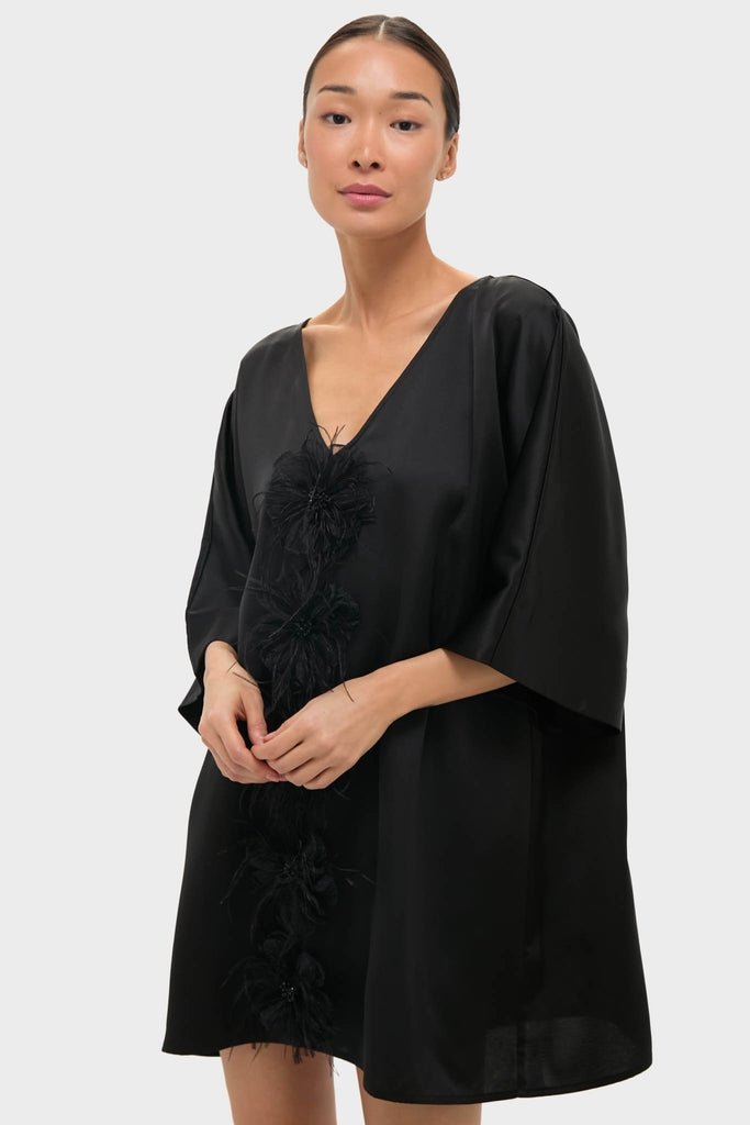 Young woman models the Black Satin Diane Mini Dress, featuring classic kimono sleeves, effortless feather embellishments, and a fun back cutout, showcasing Tuckernuck's high-end style.