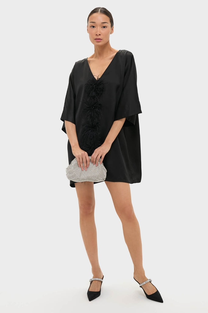 The Black Satin Diane Mini Dress features a classic V-neck, feathered rosettes, and kimono sleeves, paired with a silver clutch and sparkly slingback flats for an effortless, fun, high-end look.