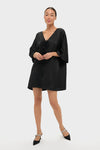 Woman models the classic Black Satin Diane Mini Dress, featuring a deep V-neck, kimono sleeves, and feather floral embellishments, embodying effortless elegance and high-end style.