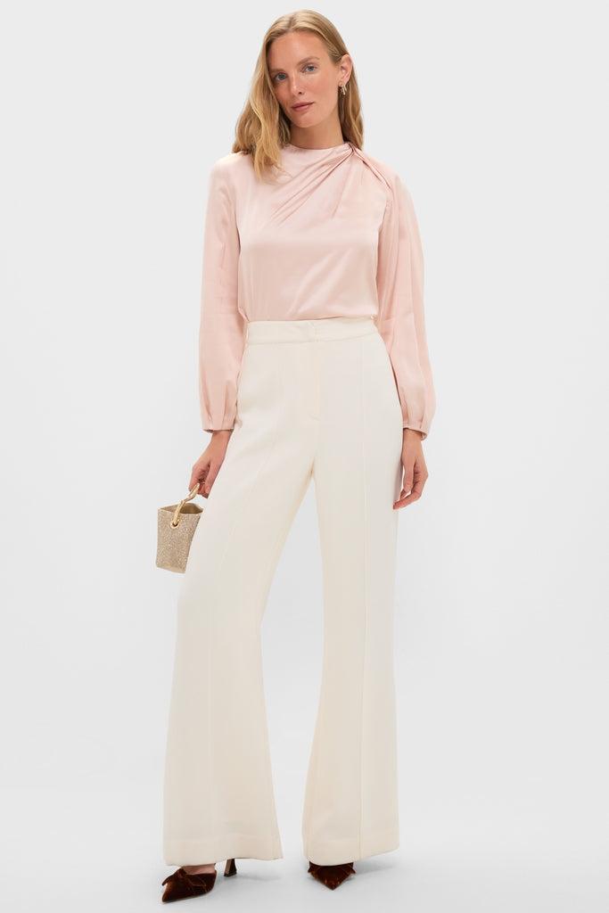 Woman in Soft Pink Blush Satin Blaire Top, featuring classic blouson sleeves and gathered shoulder, paired with cream wide-leg trousers and a fun, glittery handbag.