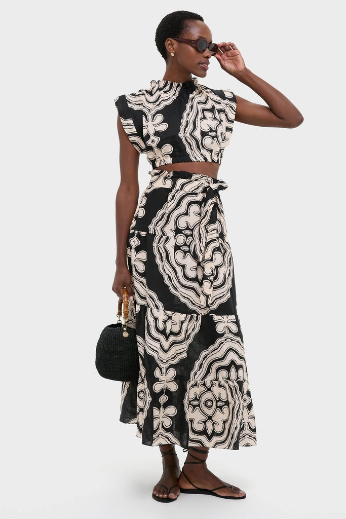 Model wears Niku Black Sanaa Skirt, featuring an effortless, high-end graphic print with a classic tie waist. Paired with matching top, black handbag, sandals, and sunglasses.