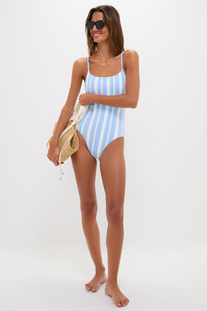 Model wearing the classic Debre Sky Elene One Piece swimsuit with effortless style, showcasing adjustable straps and high-end design, accessorized with sunglasses and a straw beach bag.
