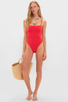 Model wearing Ruby Jenna One Piece swimsuit with square neckline, adjustable straps, and high-cut leg, holding straw tote; accessorized with necklaces and earrings, embodying effortless, high-end style.