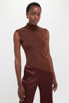 Model wearing the classic Rust Edith Top, an effortless sleeveless brown turtleneck with an asymmetrical hem, paired with high-end glossy burgundy trousers and a bold earring.