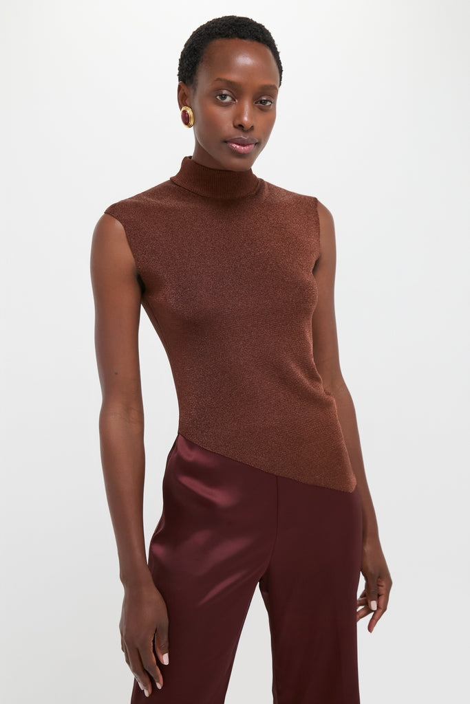 Model wearing the classic Rust Edith Top, an effortless sleeveless brown turtleneck with an asymmetrical hem, paired with high-end glossy burgundy trousers and a bold earring.