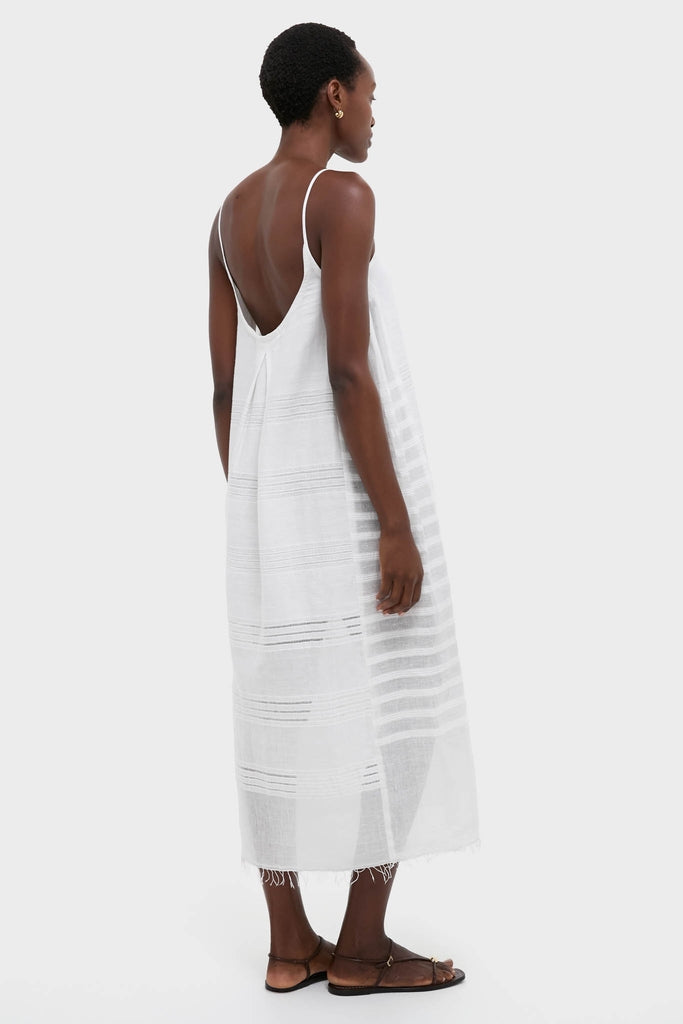 Woman models the classic Abira White Nia Slip Dress, featuring effortless sheer stripes, shimmering dots, and a hand-twisted fringe hem, paired with simple brown sandals.